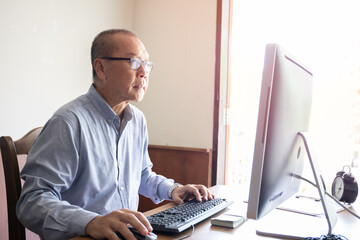 An old man using computer and looking or watching desktop monitor for business financial monitoring, elder working at home, retired man hobby
