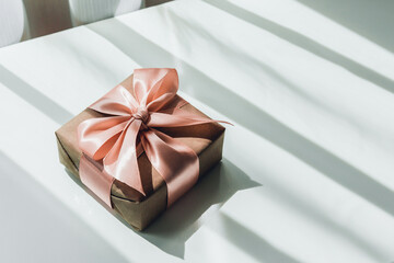 Gift box in wrapping craft paper with a pink ribbon on a white table with hard shadows.