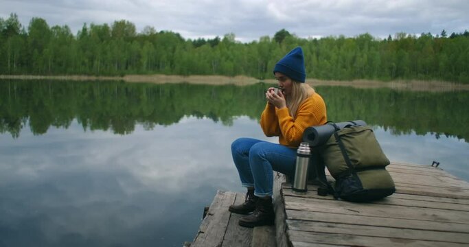 A woman with a hat and a backpack sits on a wooden pier of a forest lake in the mountains and drinks hot drinks tea or coffee from a thermos.