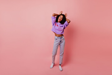 Full-length shot of inspired black girl joking during photoshoot. Carefree curly woman making funny faces on pink background.