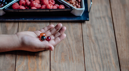 Child holds berries in the palm of his hand over a wooden table. Vases with raspberries and currants are lying on the table. Delicious food, dessert