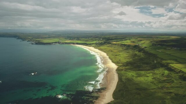 Aerial view sandy coastline, green grass meadows. Waves crashing out of shore and come back to ocean. Atlantic bay serene scenery in dusk summer daytime. Footage cinematic shot in 4K, UHD