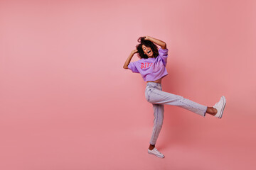 Full-length portrait of carefree slim lady in jeans. Blissful african woman dancing on rosy background.