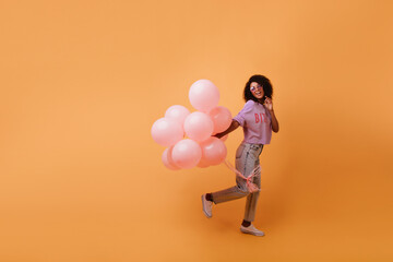 Fototapeta na wymiar Full-length portrait of charming african girl dancing on orange background. Studio photo of ecstatic black female model jumping with balloons.