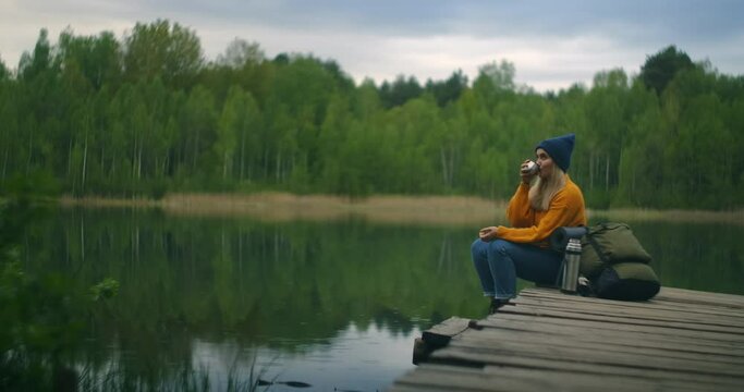 A female traveler drinks tea with a backpack and enjoys the beauty of a mountain forest lake sitting on a wooden pier. Slow-motion concept of a lonely woman's solitary journey. Quarantine and travel