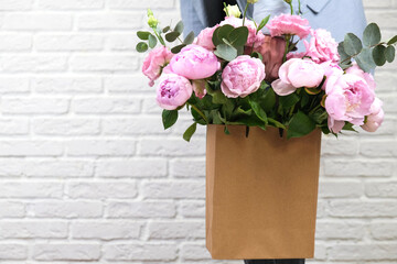 Flower delivery concept of packing flowers. The girl holds pink peonies in a craft bag, against the background of a white brick wall. Mock up.