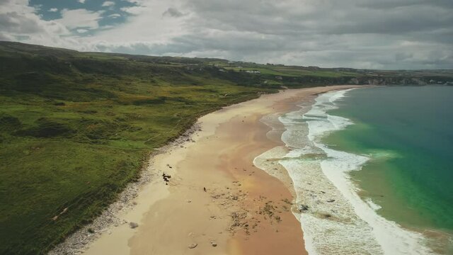 Aerial coast beach: Atlantic Ocean, Antrim county, Northern Ireland. People walking on sandy white shore with tranquil coastal wavy water. Cloudy summer scenery. Footage shot view in 4K,UHD