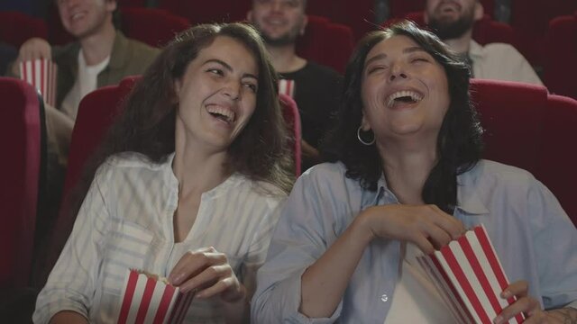 Medium shot of two attractive diverse women sitting in cinema, eating popcorn, watching comedy and laughing