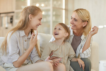 Obraz na płótnie Canvas Cute warm-toned portrait of carefree young mother talking to two daughters and smiling cheerfully while enjoying time together at home