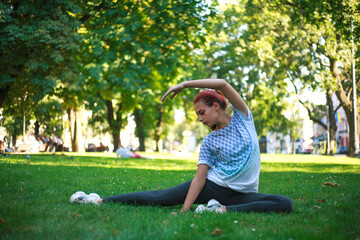 Young woman in city park on green grass doing stretching fitness exercises outdoors