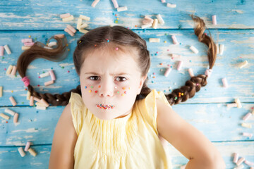 Funny emotional little girl with sprinkled lips and freckles on color background with colorful candies