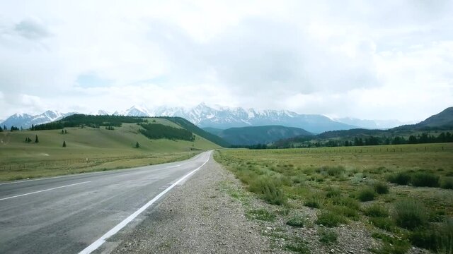 Road in the field on the background of snow-capped mountain peaks. Scene. Alps. Frontal car traveling. POV video from auto onboard mountain view on cloudy sky back.