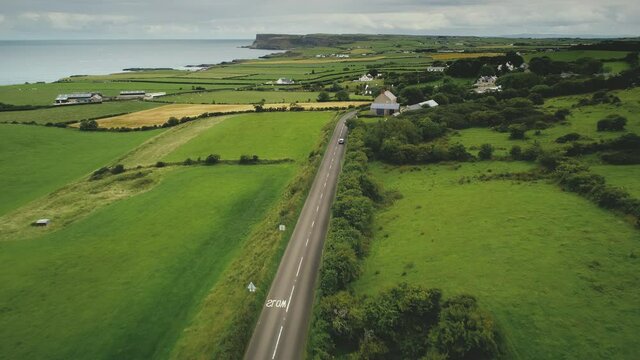 Ireland rural road aerial view: car driving up farmland. Irish green grass meadow, fields and pastures in summer evening. Majestic Irish landscape of countryside roadway. Footage shot in 4K, UHD