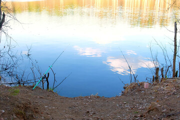 Two fishing rods on riverbank at sunset in beautiful place on nature. Catching fish concept. Clouds and trees are reflected in the water.