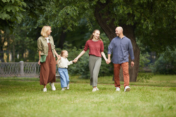 Naklejka na ściany i meble Full length portrait of modern carefree family with two kids holding hands while walking towards camera on green grass outdoors, copy space