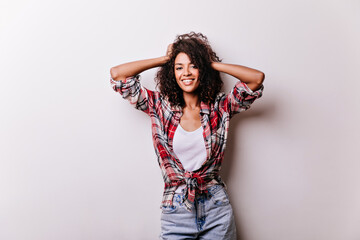 Shapely slim lady in casual attire touching her wavy hair. Appealing african girl standing on white background.