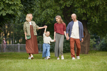 Fototapeta premium Full length portrait of carefree family with two kids holding hands while walking towards camera on green grass outdoors, copy space