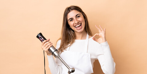 Woman using hand blender over isolated background showing ok sign with two hands