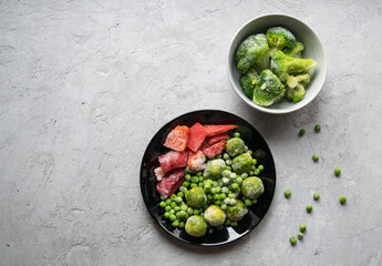 Frozen vegetables: red bell peppers, green peas, broccoli and Brussels sprouts on a gray background. Image with copy space, top view, vertical orientation.