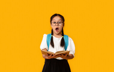 Shocked Elementary School Girl Holding Book Standing On Yellow Background