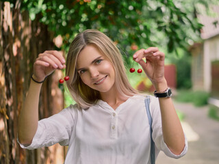 Outdoor summer city lifestyle portrait of young trendy dressed blonde happy woman having fun outdoor in the city using cherries as earrings