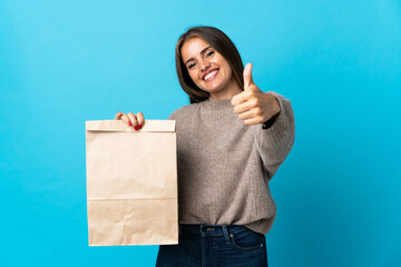 Woman taking a bag of takeaway food isolated on blue background with thumbs up because something good has happened
