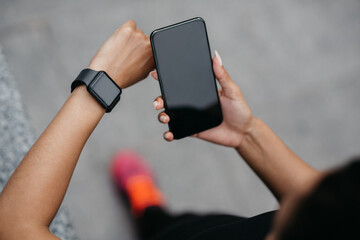 Effective training. African american girl checks smartphone and fitness tracker with empty screen on street