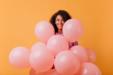 Fototapeta na wymiar Happy birthday girl posing with cheerful smile. Indoor portrait of pretty african woman with party balloons isolated on orange background.