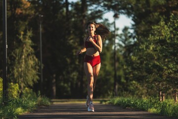 Athletic woman in a red top and shorts running in summer park on road and smiling. Green trees on the background.