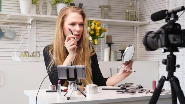 Vlogger female applies lipstick on lips. Beauty blogger woman filming daily makeup routine tutorial at camera on tripod. Influencer blonde girl live streaming cosmetics product review in home studio