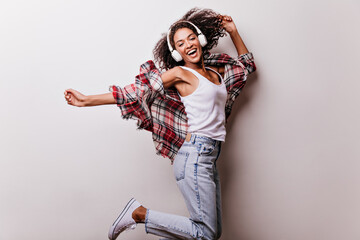 Cute curly girl in jeans jumping with happy face expression. Good-humoured african lady in headphones expressing positive emotions during photoshoot on white background.