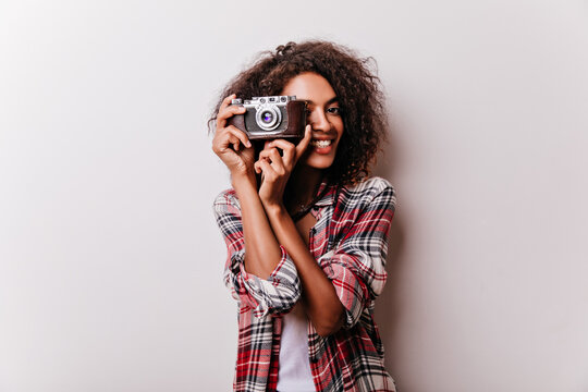 Blissful smiling girl with camera making photos. Indoor shot of attractive female photographer wears checkered shirt.