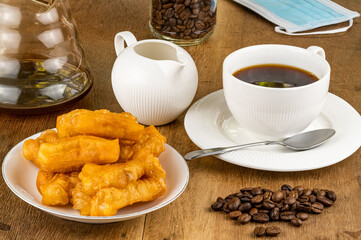 Breakfast with deep-fried dough stick in a white ceramic plate and black coffee in a white ceramic cup with a pile of coffee beans on a wooden table.