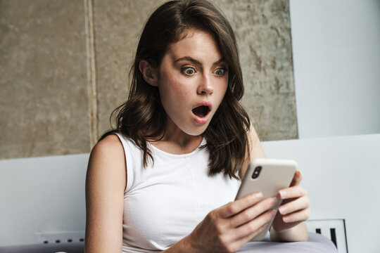 Image of brunette shocked woman using smartphone while resting on couch