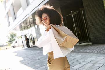 Attractive young African-American woman walks on a shopping spree in the city. Beautiful trendy girl carries paper shopping bags and looks back with a smile