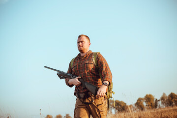 Strong young hunter with red beard holding his gun and walking along the dirt road under blue sky