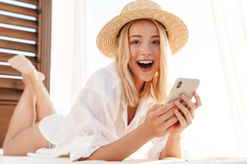 Image of excited blonde woman using cellphone while lying on bed