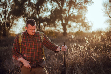 Naklejka na ściany i meble Irish looking red bearded hunter in checkered shirt staying outdoors in the sunset light and holding his gun