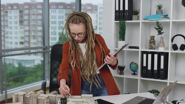 Pretty concentrated busy smart stylish young woman with dreadlocks doing notes into folder about technical construction of new buildings in design studio,4k