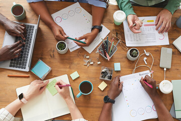 Top view close up of multi-ethnic group of people working together at cluttered wooden table with coffee cups, mugs and stationary items, teamworking or studying concept, copy space - obrazy, fototapety, plakaty