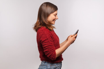 Side view, happy brunette girl in shaggy sweater using mobile phone and smiling laughing joyfully,...