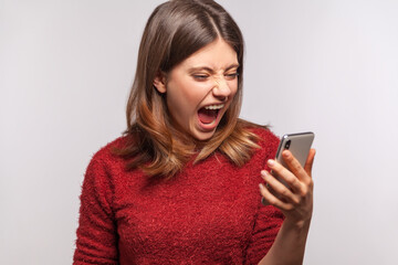 Fototapeta premium Angry irritated woman keeping mouth open roaring wild, shouting while looking at cell phone screen with crazy furious expression, aggressive mobile user. indoor studio shot isolated on gray background