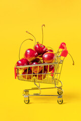 Metal shopping trolley filled with organic sweet cherry berries on yellow background. Shopping...