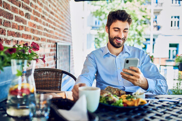 Fototapeta na wymiar Cheerful young entrepreneur eating breakfast in outdoor cafe and using smartphone