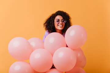 Pleased young woman in round glasses posing with pink balloons. Indoor photo of joyful african birthday girl isolated on yellow background.