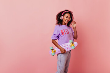 Cute female black model holding purple skateboard. Adorable african woman with curly hairstyle listening favorite song and smiling.