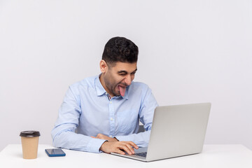 Funny man employee sitting office workplace with laptop on desk, showing his tongue out while talking video call on laptop, disobedient behavior. indoor studio shot isolated on white background