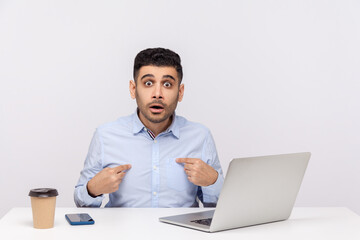 Unbelievable, this is me! Amazed businessman sitting office workplace with laptop on desk, pointing himself and looking at camera with surprised shocked expression. indoor studio shot isolated