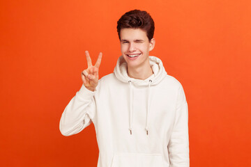 Happy positive teenager in stylish sweatshirt showing v sign to camera, winking and smiling, young...