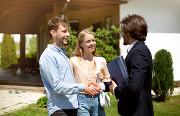 Young married couple shaking hands with realtor, making deal about house purchase outside